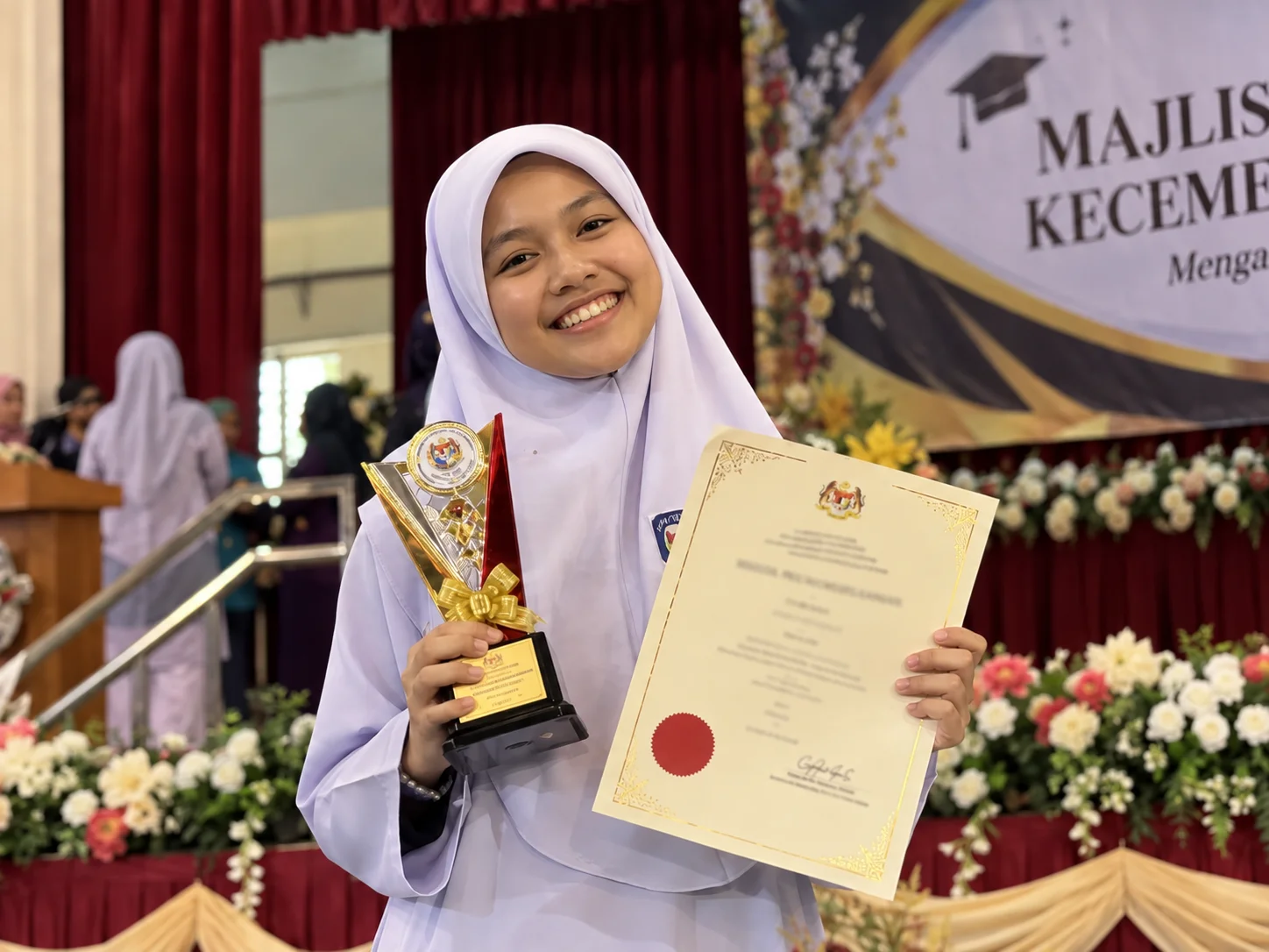 Pelajar cemerlang tersenyum gembira memegang piala anugerah dan sijil kejayaan di sekolah